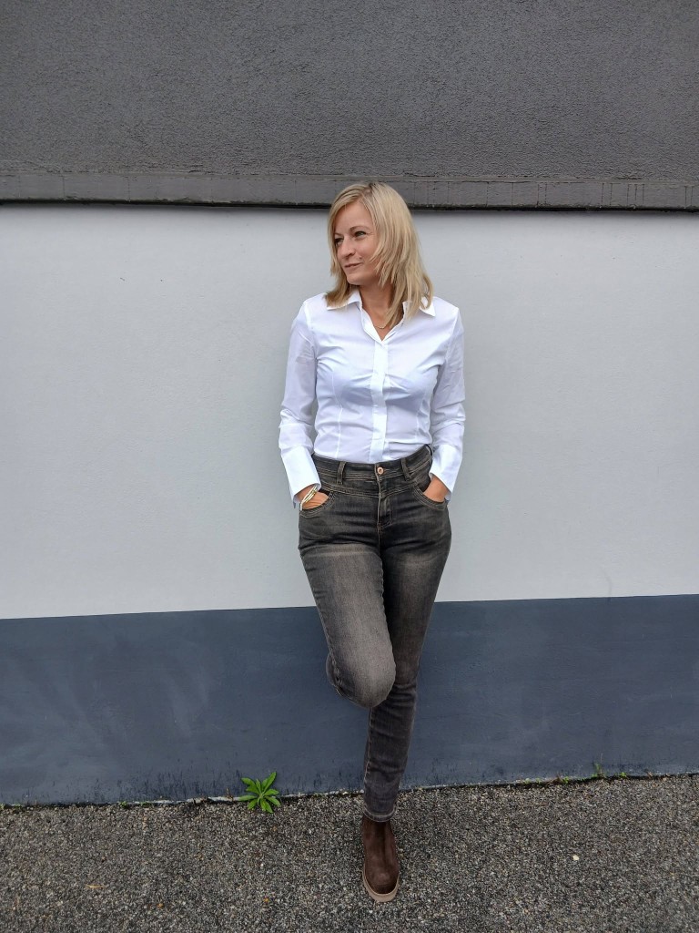 Laurie en chemise blanche et jean sombre, adossée au mur, une jambe croisée